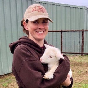 A woman smiles at the camera while holding a baby lamb in her arms
