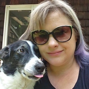 A woman in sunglasses smiles at the camera as her dog looks lovingly at her