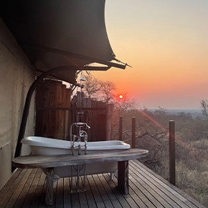 The side of a building with bath facilities overlooks the sun setting over brush in the savanna.
