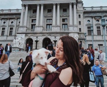 A young woman holding a white dog and standing in front of a government building.