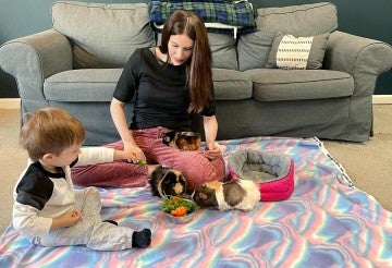 A woman and her young son play with three guinea pigs on the floor while an inset image shows one of the guinea pigs in the filthy conditions it was rescued from before adoption