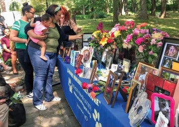 People grieving in a park with a table full of flowers and pet photos.