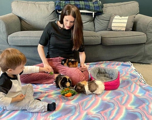 A woman and her young son play with three guinea pigs on the floor while an inset image shows one of the guinea pigs in the filthy conditions it was rescued from before adoption