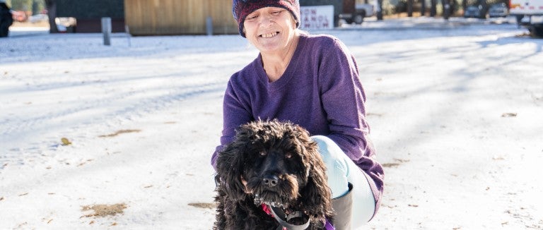A woman in a purple beanie and shirt holds a black, curly haired dog in the snow